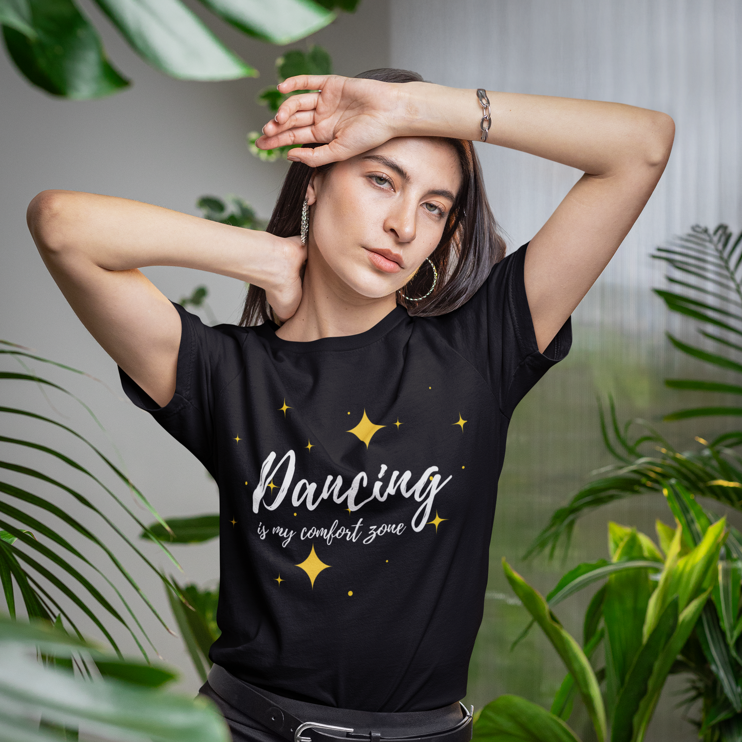 Woman wearing a black unisex dance t-shirt with 'Dancing is my comfort zone' text, surrounded by green plants.