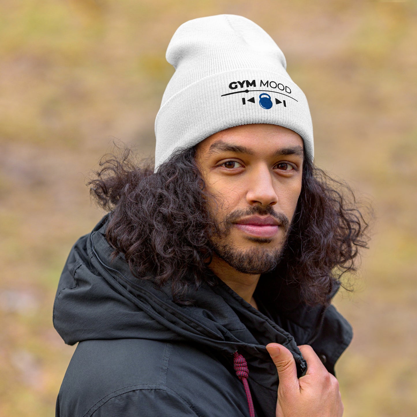 Man wearing a white Gym Mood On beanie with black text and blue kettlebell embroidery, outdoors with a blurred natural background.
