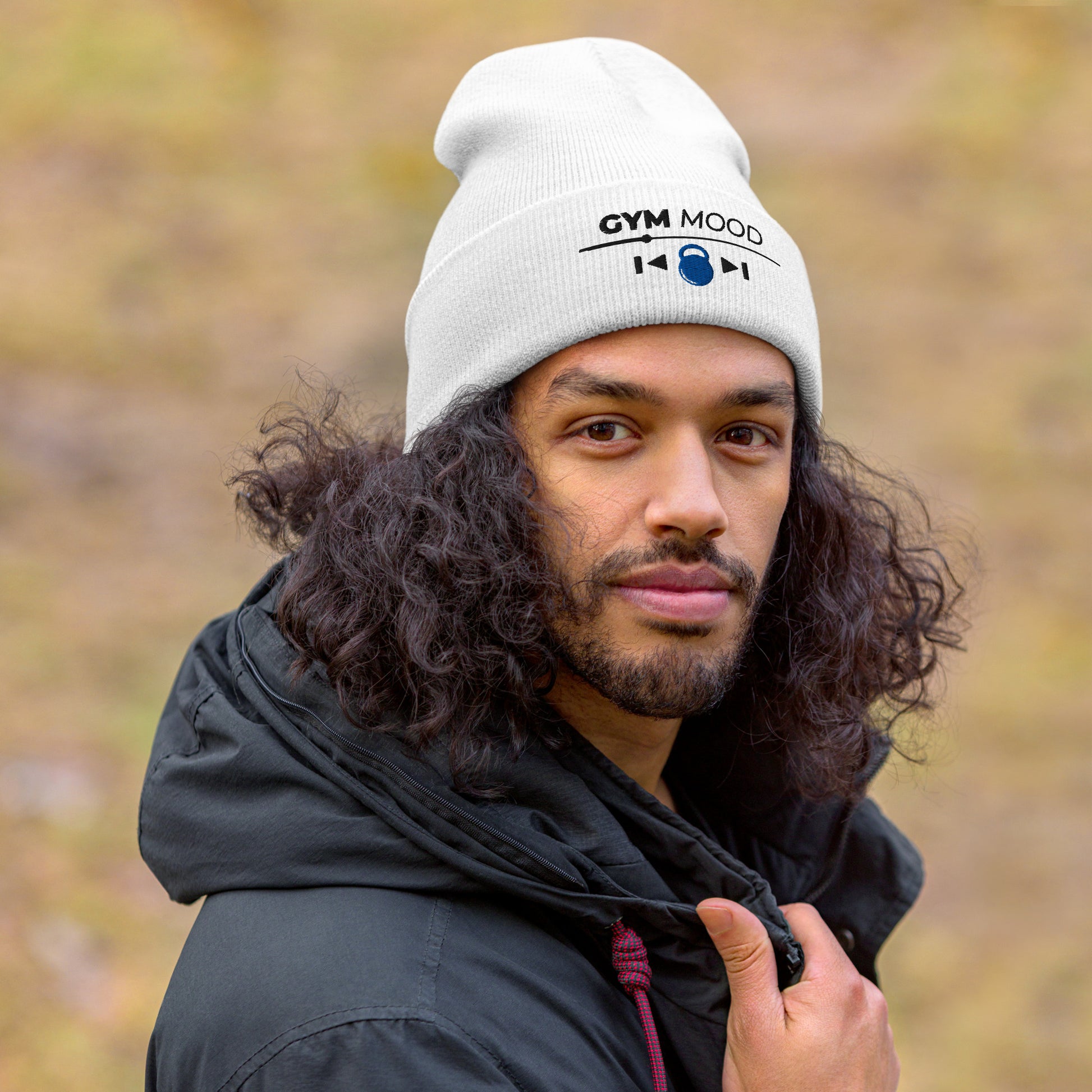 Man wearing a white Gym Mood On beanie with black text and blue kettlebell embroidery, outdoors with a blurred natural background.