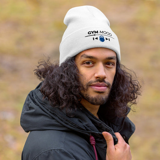 Man wearing a white Gym Mood On beanie with black text and blue kettlebell embroidery, outdoors with a blurred natural background.