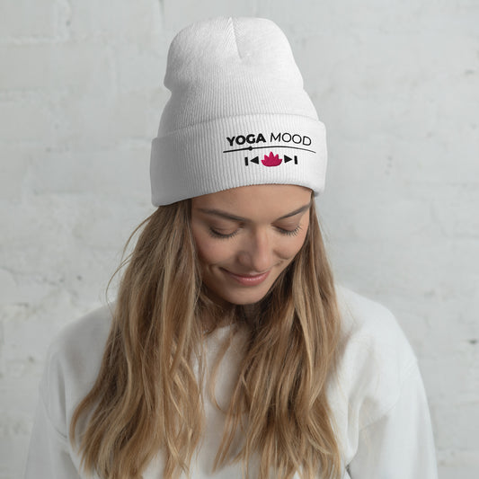 Woman wearing a white Yoga Mood On beanie with black text and pink lotus embroidery, smiling against a light gray background.