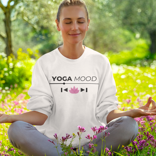 White unisex Yoga Mood On sweatshirt featuring black text and pink lotus graphic, worn by a woman sitting cross-legged in a flower field.